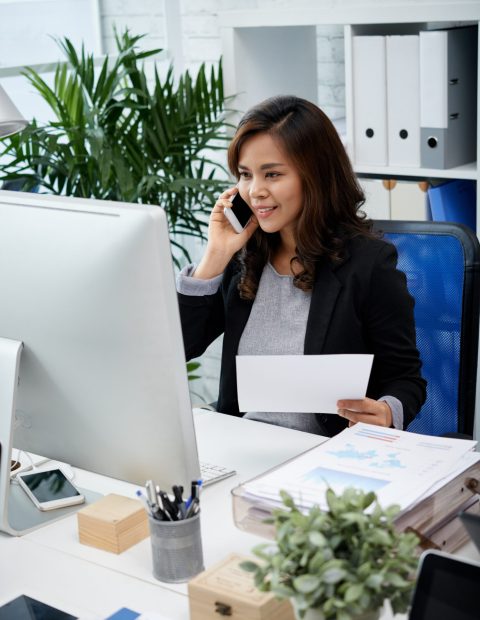 Pretty female business executive sitting at her table and calling on the phone to her colleague